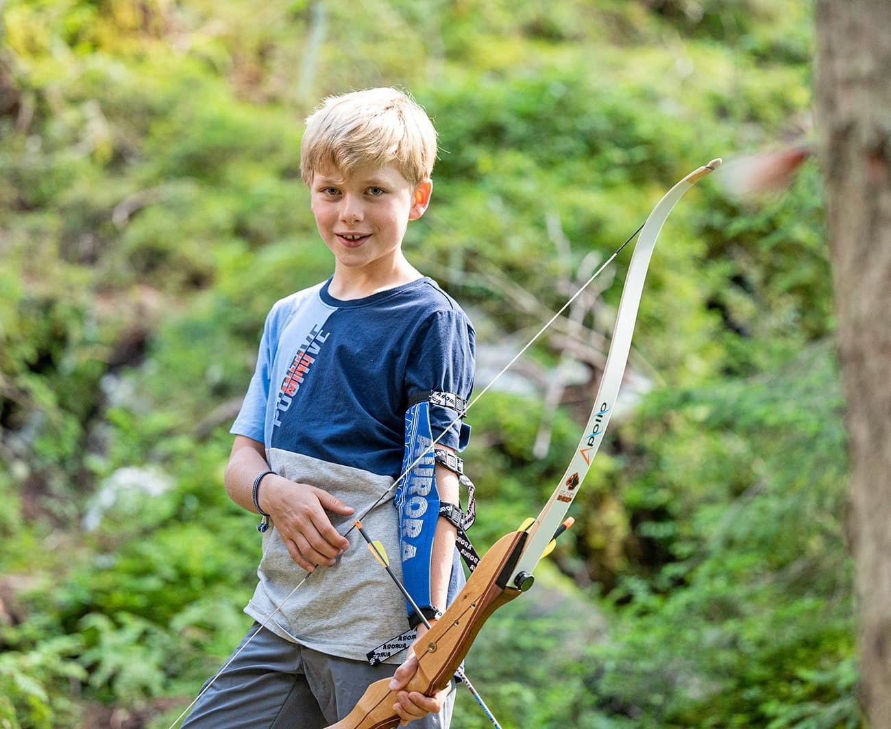 A young boy holding a bow and arrow stands in a lush, green forest