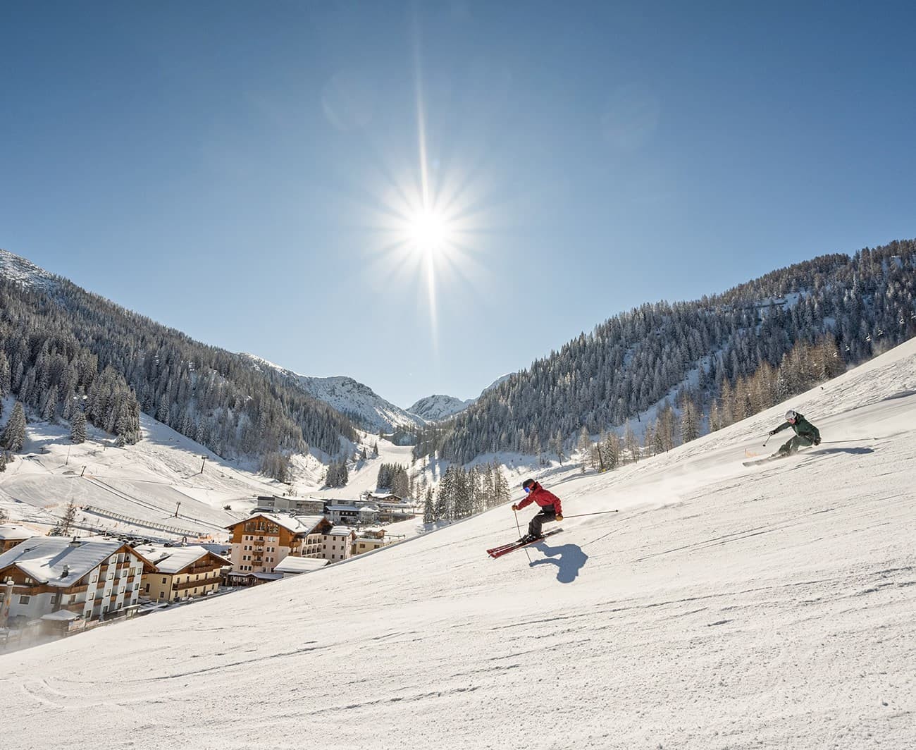 Two skiers descend a snowy slope under a bright sun and clear blue sky in Zauchensee