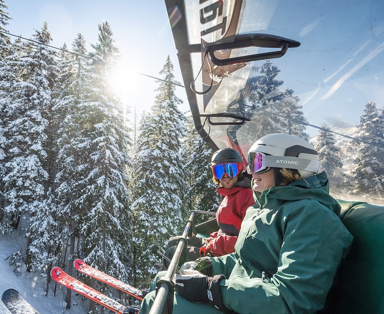 Two skiers on a lift, surrounded by snowy trees under a bright sun
