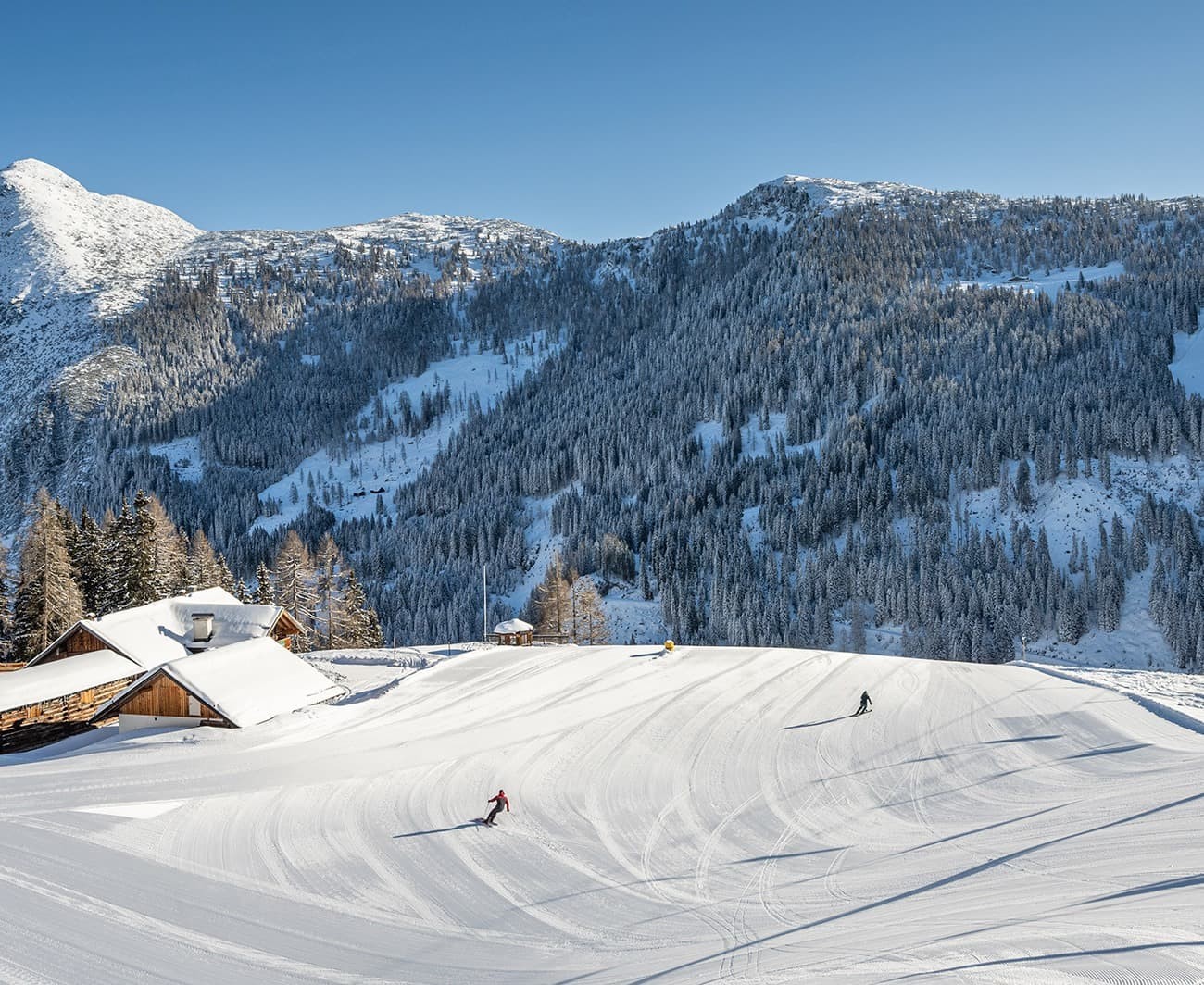Skiers on a snowy slope near a cabin with mountain backdrop under a clear blue sky