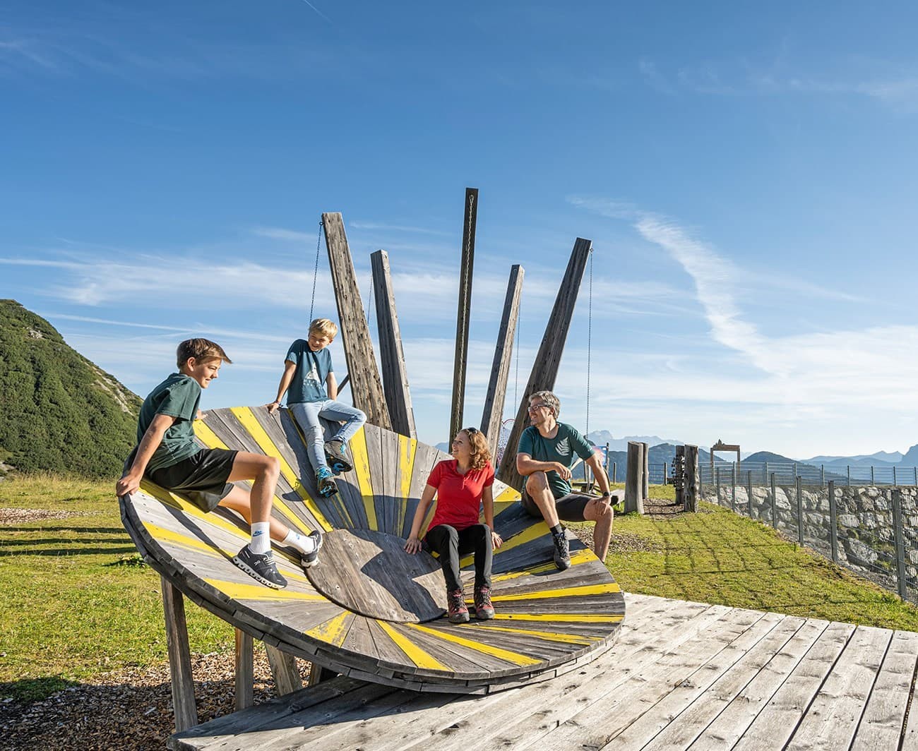 Four people sit and chat on a wooden spiral sculpture in a mountainous landscape
