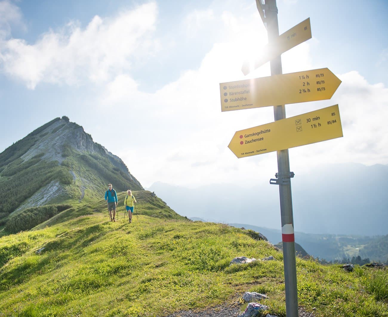 Two hikers near a mountain path with direction signs under a bright sky