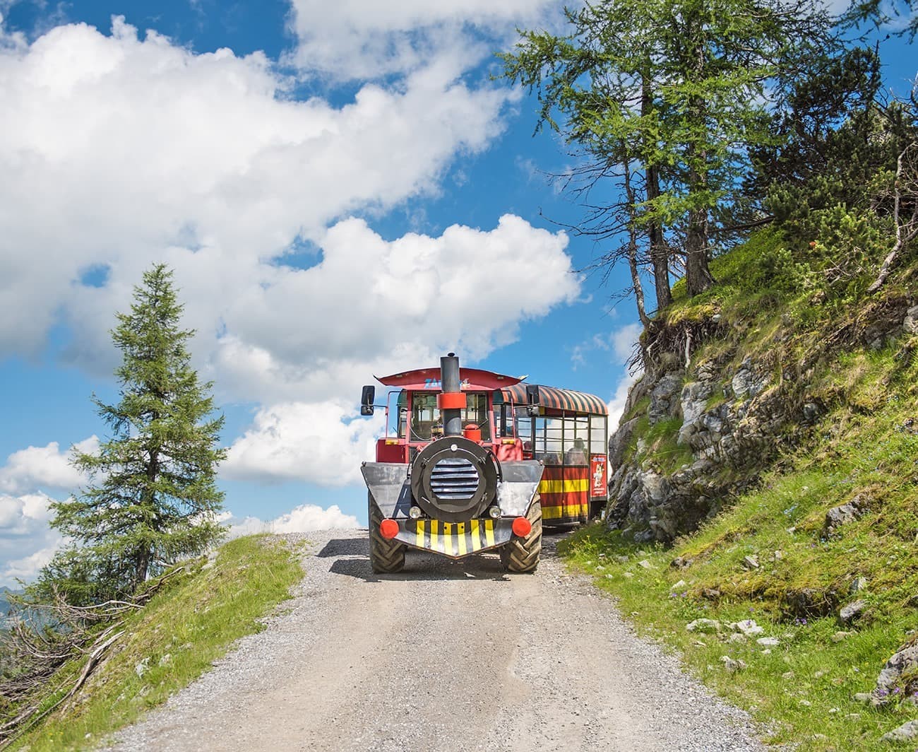 A colorful tourist train travels on a scenic mountain path under a blue sky